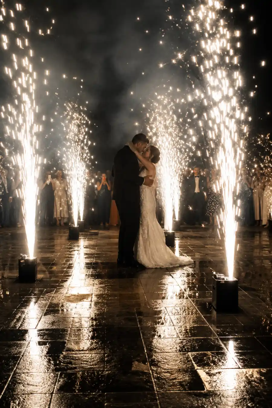 Newlyweds watch pale and gilt fireworks fill the night sky over a wet stone terrace at their wedding celebration at midnight