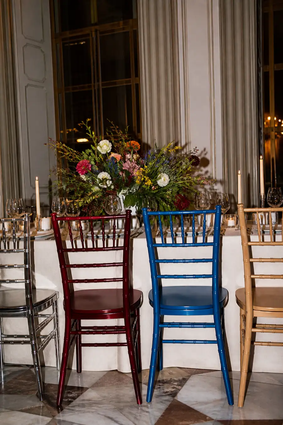 Silver, oxblood, cobalt and natural wood chairs surround a candlelit wedding table topped with a colorful floral centerpiece.