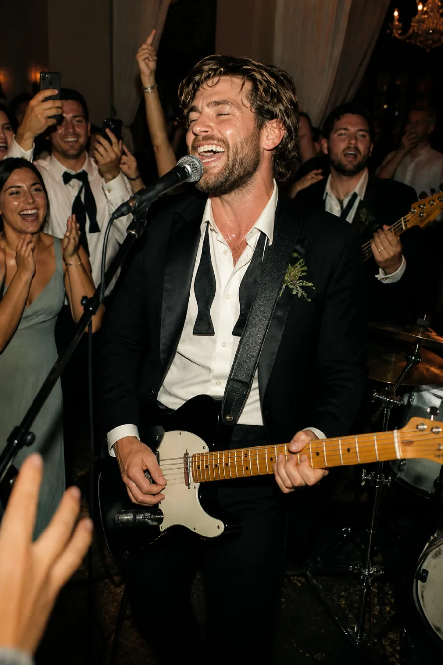 The groom sings and plays electric guitar on the reception dance floor while friends and musicians cheer around him with joy.