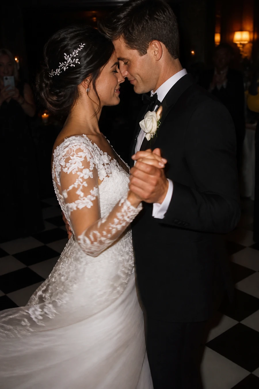Newlyweds share an intimate first dance on a checkerboard floor while warm lamps glow around the dark reception room at dusk.