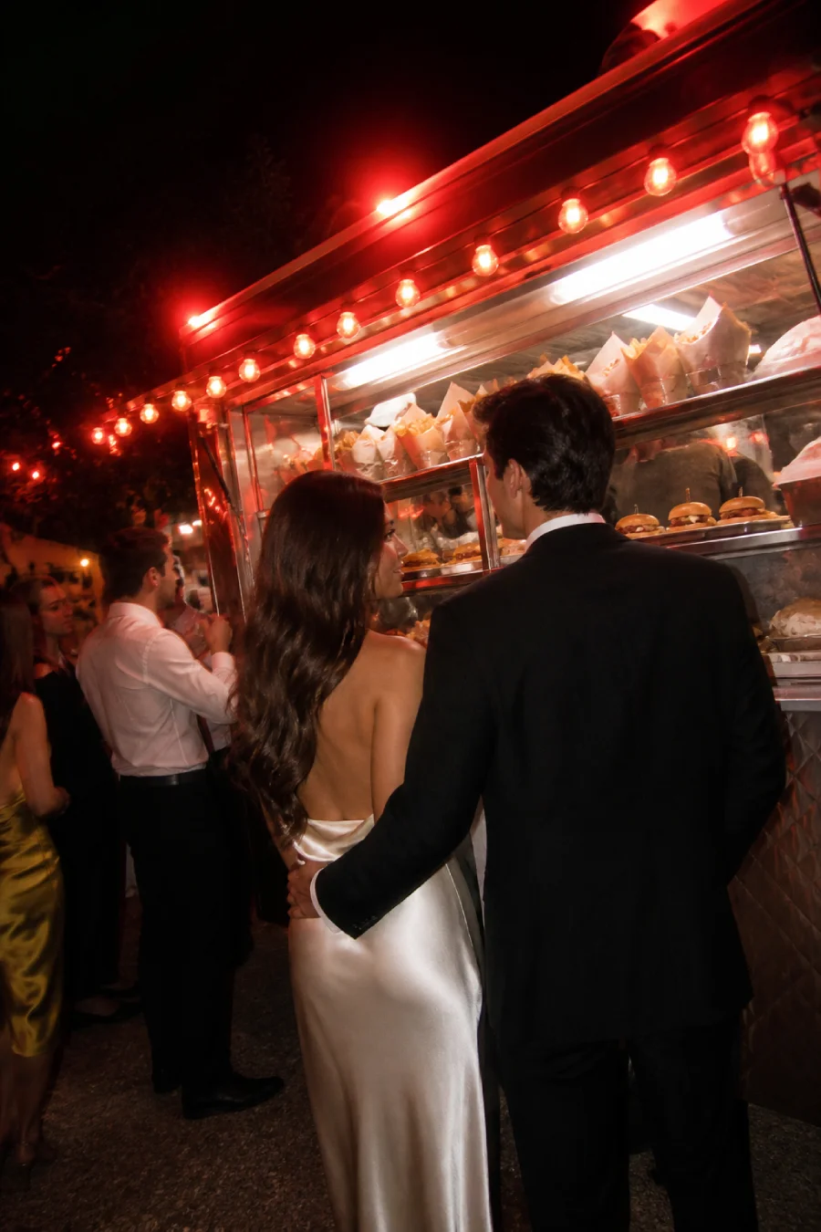 Newlyweds wait beside a silver late-night food truck serving fries and sliders under vivid ruby lights after their reception.