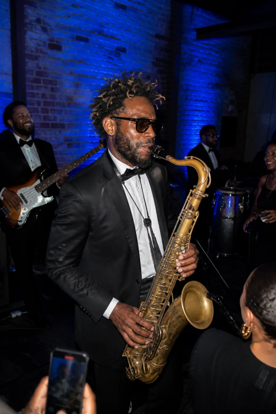 Saxophonist performs with a live band under cobalt lighting as wedding guests gather close to the stage inside the reception