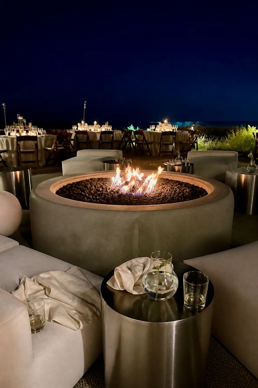 Round firepit glows between ivory lounge seats and silver tables beside candlelit wedding tables overlooking the ocean below