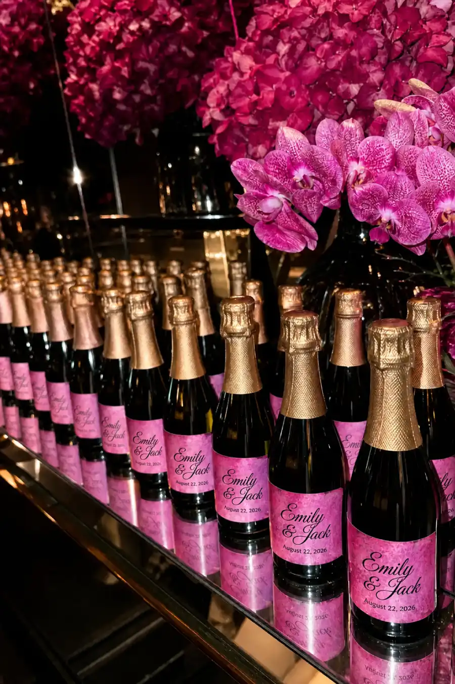 Personalized champagne bottles with pink labels and gold lettering beside vibrant magenta wedding flowers
