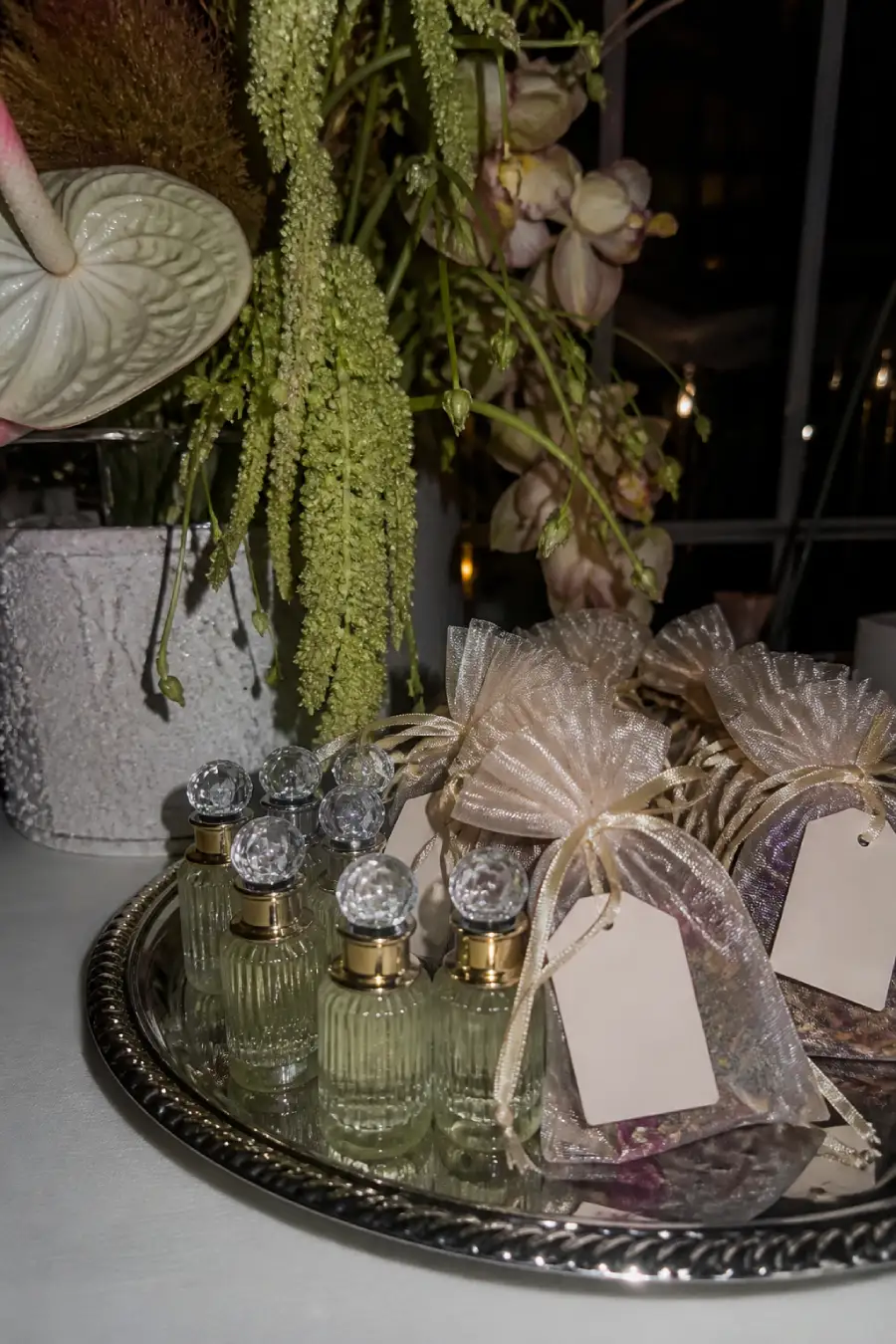 Elegant glossy perfume bottles and translucent scent sachets rest on a silver tray beneath sculptural wedding flowers nearby