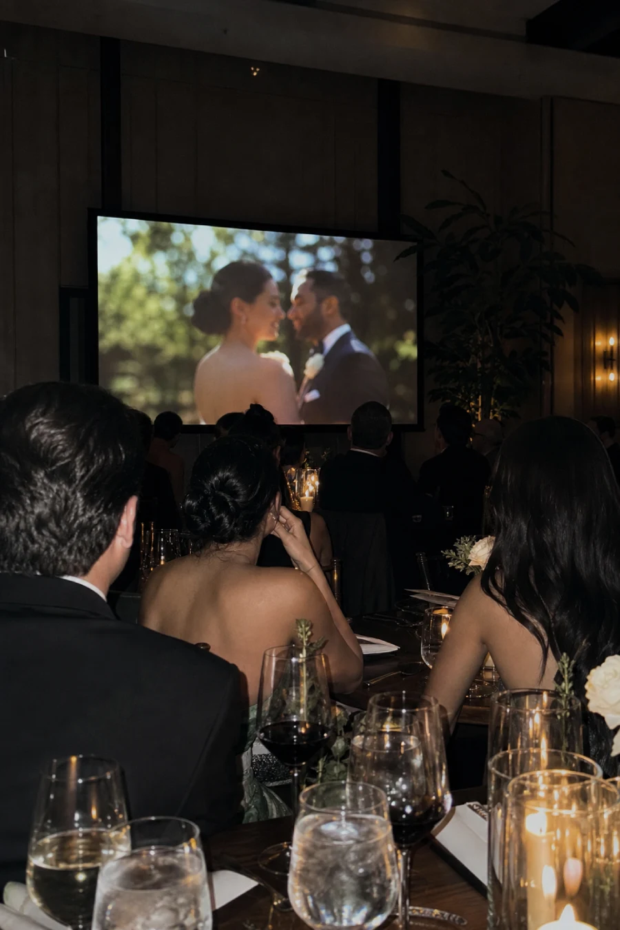 Wedding guests watch the couple's love story video from candlelit dinner tables inside a dim contemporary reception ballroom.