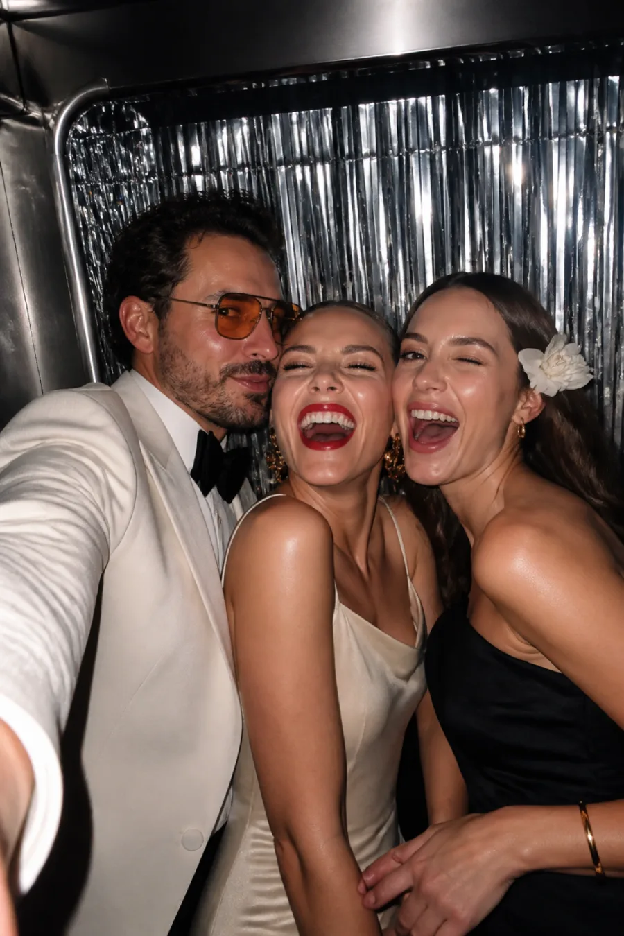 Three fashionable wedding guests laugh together inside the silver-fringed photo booth while posing for a direct-flash selfie