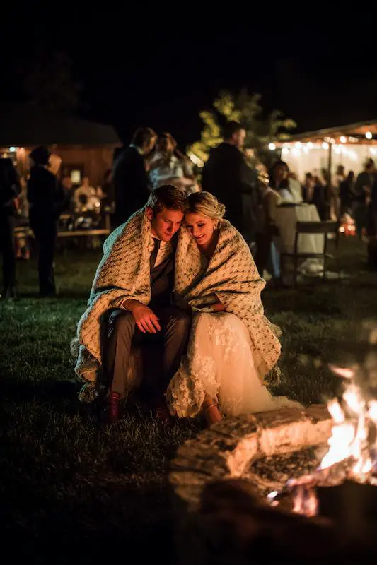 Bride and groom wrapped in a blanket share a cozy moment by the fire pit during their outdoor night wedding reception. Bride and groom wrapped in a blanket share a cozy moment by the fire pit during their outdoor night wedding reception.