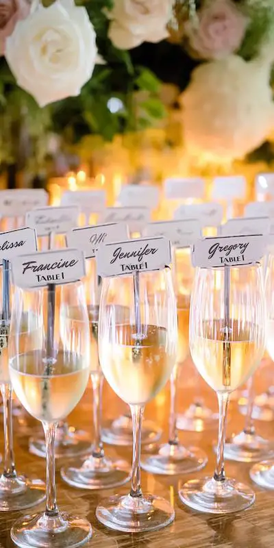 Champagne glasses with escort cards displaying guest names and table numbers, set against candles and flowers. Champagne glasses with escort cards displaying guest names and table numbers, set against candles and flowers.