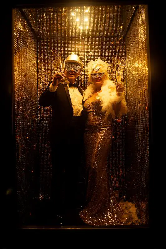Couple in glamorous attire posing in a gold sequin photo booth, wearing playful masks and holding champagne glasses, creating a festive and celebratory vibe Couple in glamorous attire posing in a gold sequin photo booth, wearing playful masks and holding champagne glasses, creating a festive and celebratory vibe