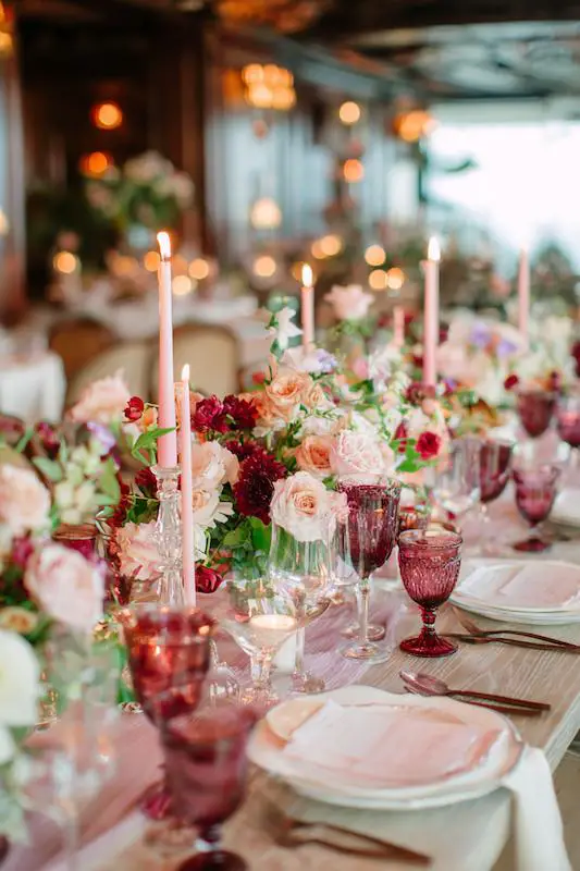 Elegant wedding reception table with pink taper candles, lush floral centerpieces of roses and greenery, and vintage-style glassware in shades of blush and berry. Wedding Reception Ideas in the form of an Elegant wedding reception table with pink taper candles, lush floral centerpieces of roses and greenery, and vintage-style glassware in shades of blush and berry.