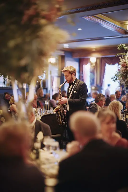 Saxophonist in a tuxedo performing live during a black-tie wedding reception, surrounded by elegantly dressed guests seated at candlelit dinner tables Saxophonist in a tuxedo performing live during a black-tie wedding reception, surrounded by elegantly dressed guests seated at candlelit dinner tables