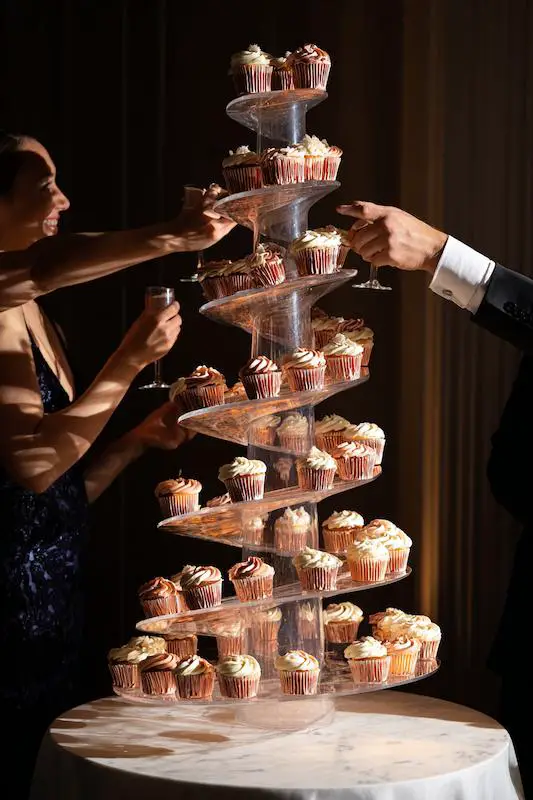 Towering spiral cupcake display at a wedding, with guests reaching for cupcakes while holding champagne glasses, lit dramatically for a modern dessert table presentation Towering spiral cupcake display at a wedding, with guests reaching for cupcakes while holding champagne glasses, lit dramatically for a modern dessert table presentation