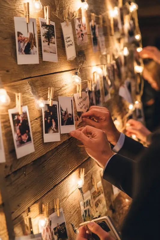 Wedding guest pinning Polaroid photos to a wooden board with string lights as a creative guestbook alternative. Wedding guest pinning Polaroid photos to a wooden board with string lights as a creative guestbook alternative.