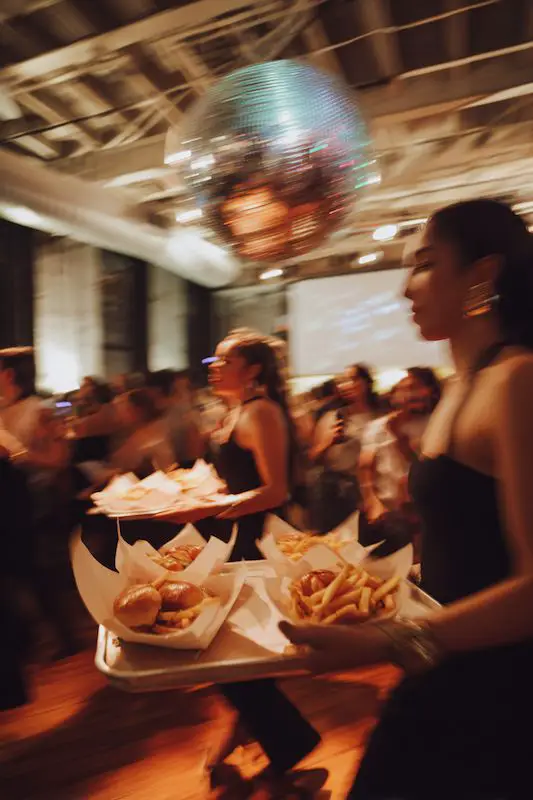 Wedding servers carrying trays of late-night sliders and fries across a lively dance floor under a disco ball. Wedding servers carrying trays of late-night sliders and fries across a lively dance floor under a disco ball.