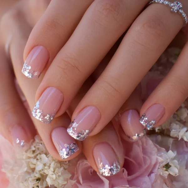 Close-up of hands with pink nails featuring silver glitter tips, resting on a bouquet of pale pink and white flowers