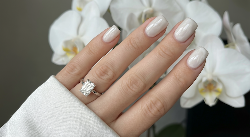 Hand with White Onyx bridal manicure showing emerald cut engagement ring in front of white orchids