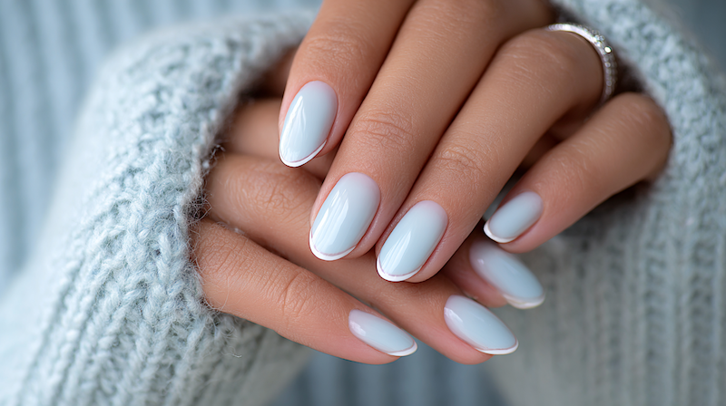 Macro close-up of hands with soft icy blue almond nails, glossy finish, subtle white outline tips, cozy knit sweater, diamond ring