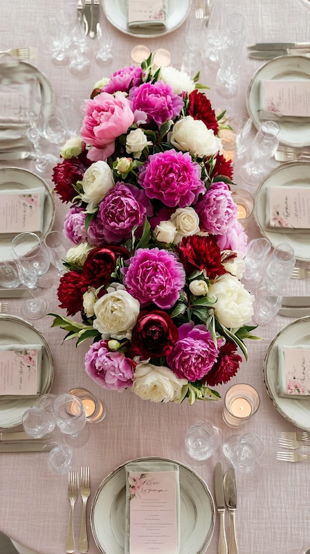 Overhead wedding tablescape with lush peony runner in pink, ivory, and burgundy with elegant place settings and soft candlelight glow