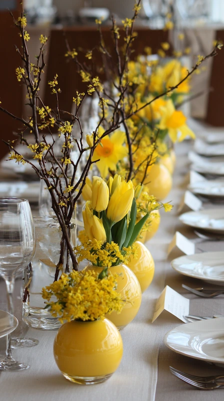 Sunny yellow wedding table with tulips and forsythia branches in round vases, white plates, place cards, and airy spring styling