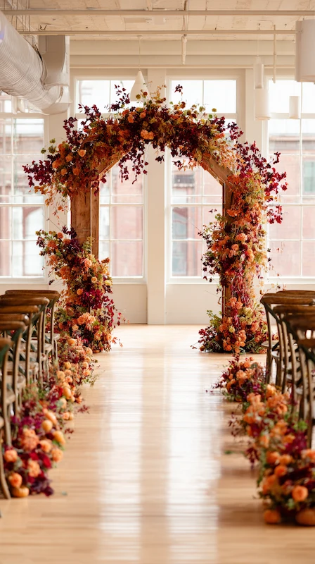 39 Wedding Arch Ideas That Will Make Your Ceremony Unforgettabl 32 Autumn wedding ceremony aisle with warm fall florals, wooden arch, and rustic chairs in a bright loft with large windows.