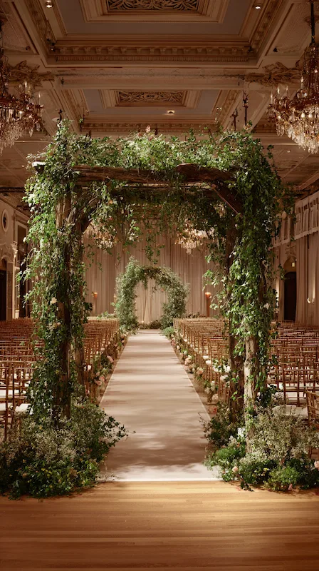 39 Wedding Arch Ideas That Will Make Your Ceremony Unforgettabl 12 Elegant indoor wedding ceremony aisle with greenery-covered wooden arch, garden florals, and rows of chairs in a grand ballroom.