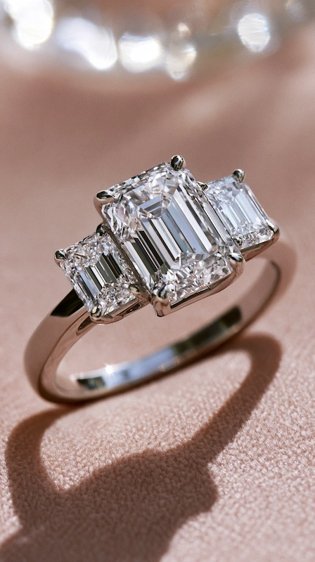 Emerald cut three stone diamond engagement ring on soft blush fabric, featuring trapezoid side stones and sleek polished band