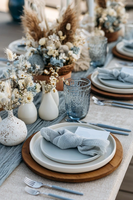 Boho dusty blue wedding tablescape with dried florals, wood chargers, textured linens, and soft neutral reception styling.