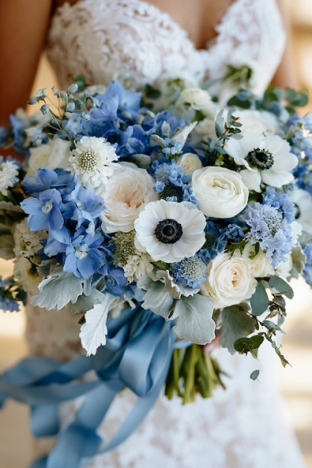Close-up of a dusty blue bridal bouquet with white roses, anemones, delphiniums, eucalyptus, and a blue silk ribbon.