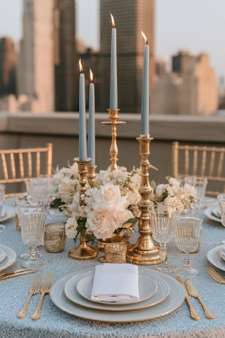 Dusty blue taper candles in brass candlesticks with white floral centerpiece on an elegant rooftop wedding reception table.