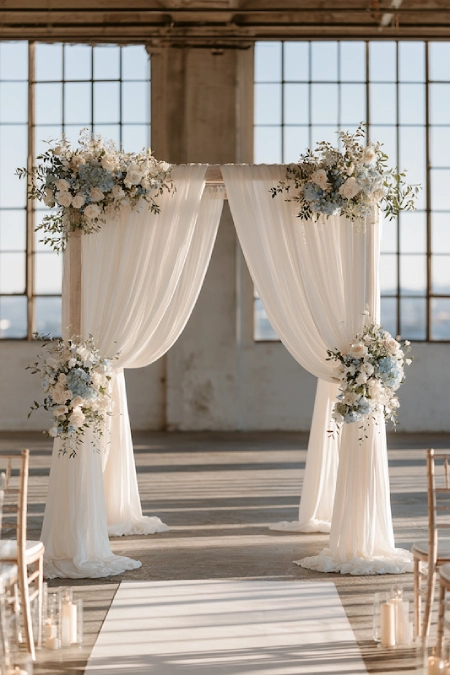 Dusty blue wedding ceremony backdrop with flowing ivory drapes, soft florals, candlelight, and a bright industrial venue.