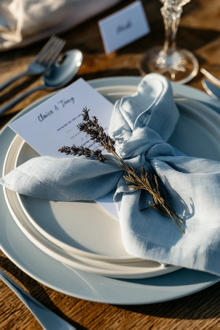 Dusty blue wedding place setting with layered white plates, linen napkin, lavender sprig, printed menu, and crystal stemware.