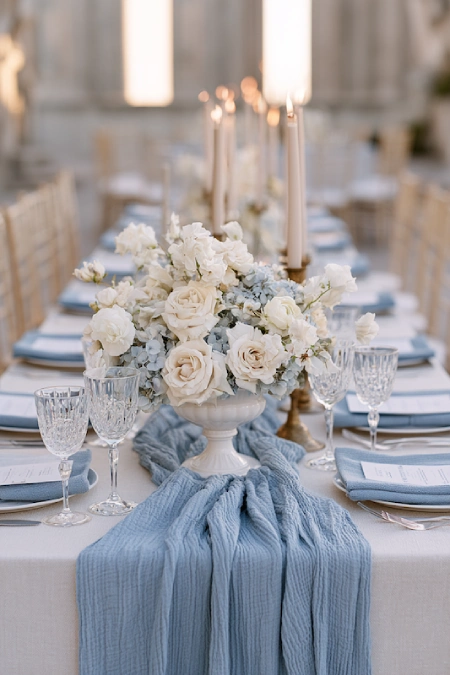Elegant wedding tablescape with dusty blue runner, white floral centerpiece, crystal glassware, and taper candles on a banquet table.