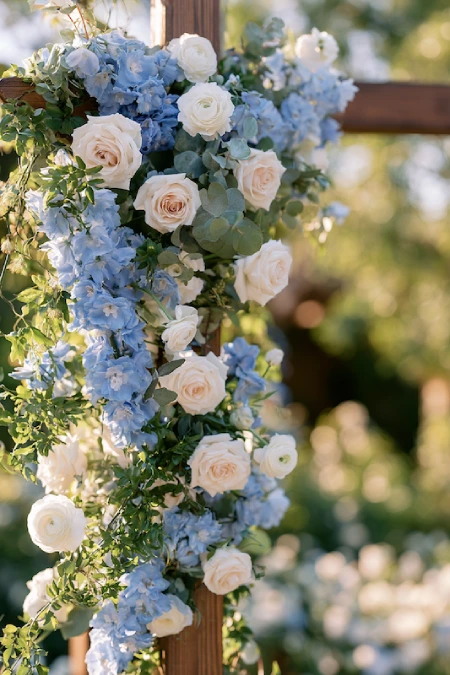 Dusty blue and white floral wedding arch with cascading roses, hydrangeas, eucalyptus, and romantic garden ceremony décor.