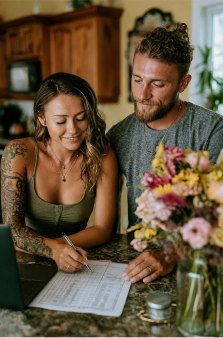 Why Weddings Go Over Budget (And Exactly Where the Money Goes) 2 Engaged couple reviewing wedding planning documents at home with flowers, laptop, and checklist on kitchen counter.