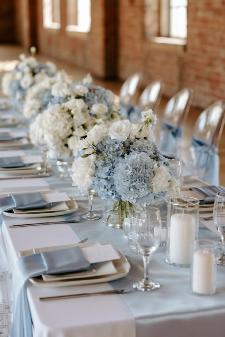 Minimalist dusty blue wedding tablescape with hydrangea centerpieces, white candles, blue napkins, and elegant reception décor.