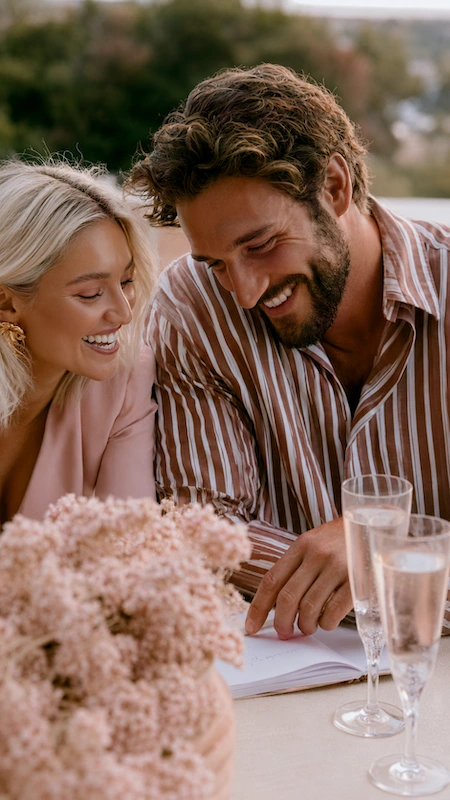What to Do First After Getting Engaged (Complete Checklist) 13 Newly engaged couple reviewing wedding planning notes with champagne and blush flowers, sharing a joyful moment together.