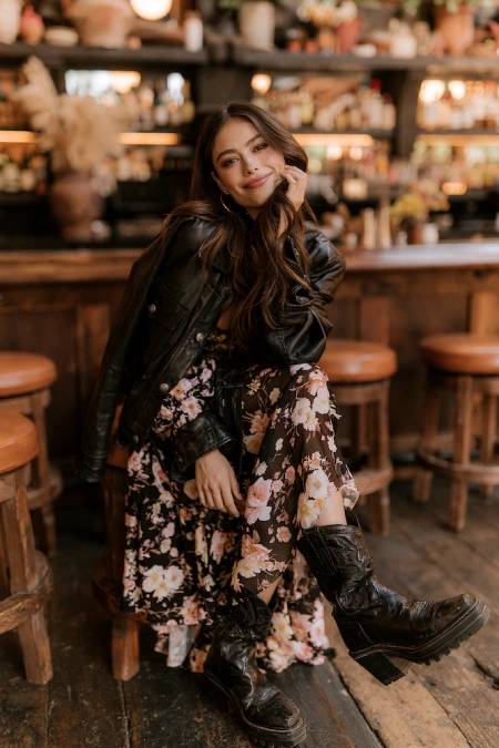 The Perfect Rehearsal Dinner Dress for Guests: Your Complete Outfit Guide 11 Woman in a floral maxi dress with a leather jacket and boots at a casual rehearsal dinner in a cozy restaurant setting