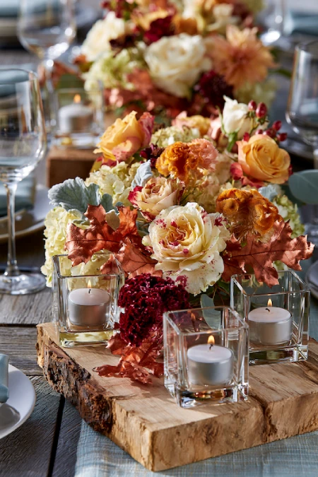 25 Fall Wedding Centerpieces With Main Character Energy (Cozy, Never Costume-y) 2 Fall wedding centerpiece with cream roses, rust florals, autumn leaves, candles, and wood risers on a rustic reception table