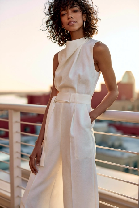 Rehearsal Dinner Dresses for the Bride: What to Wear at Every Formality Level 6 Bride-to-be in a sleek ivory jumpsuit with high neckline and belted waist posing on a rooftop terrace at golden hour