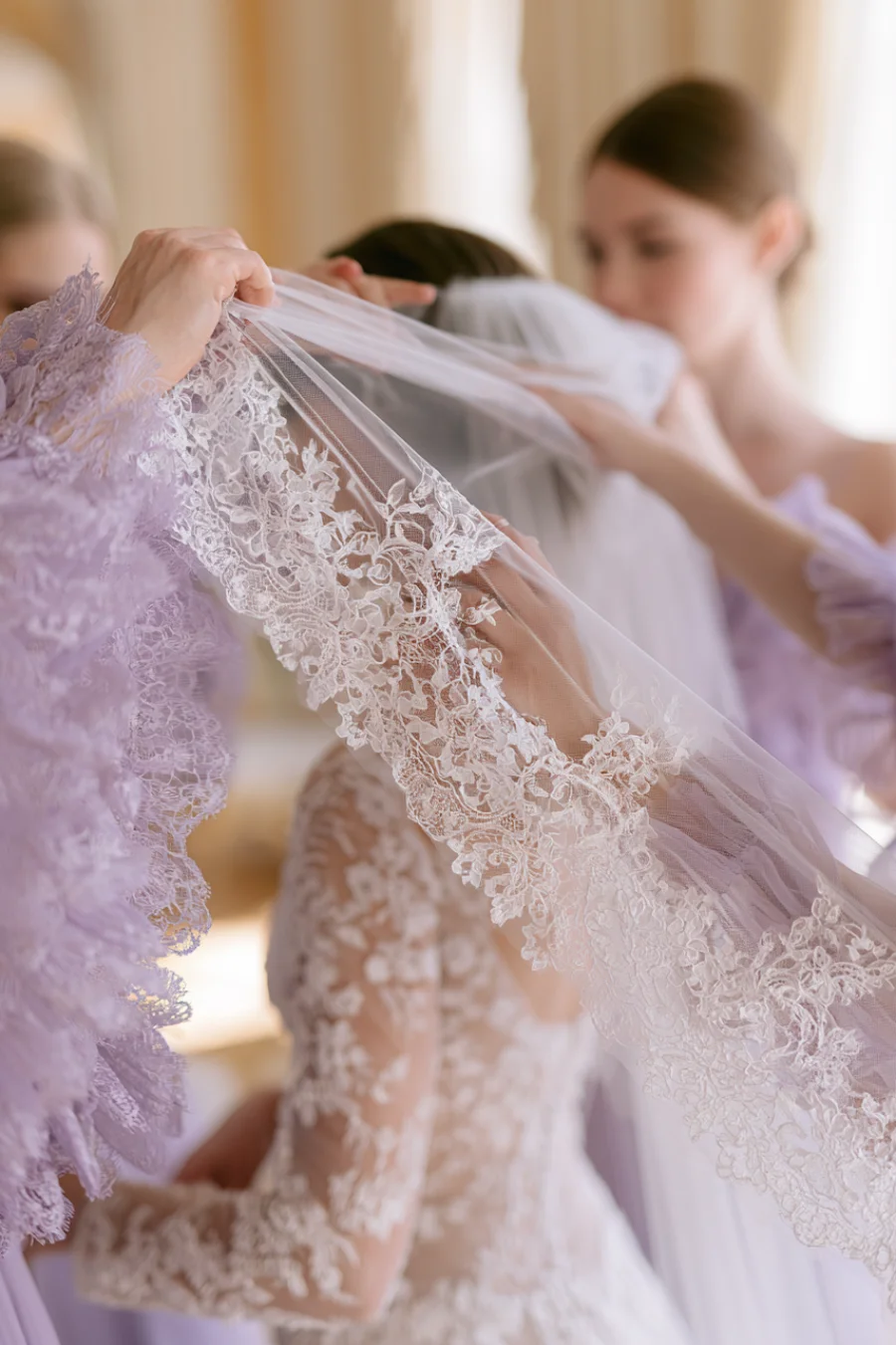 Bride wearing a lace wedding dress as attendants place a lace-edged veil over her during elegant bridal wedding preparations