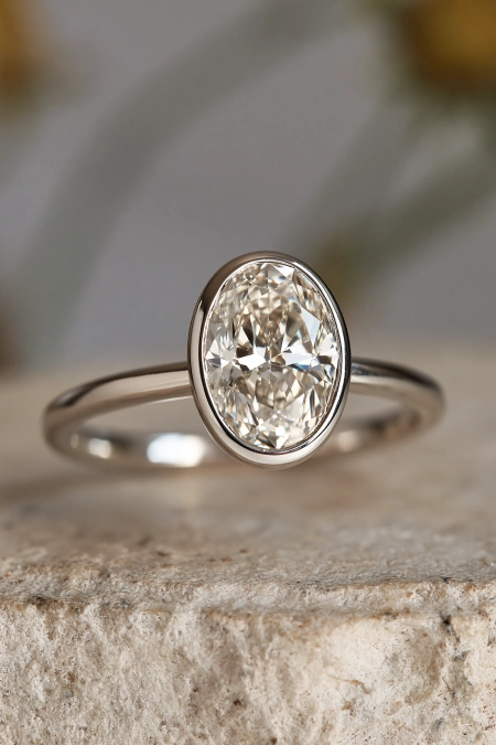 Oval Engagement Ring Obsession: Why This Cut Is Everyone's Main Character Right Now 12 Oval bezel set diamond engagement ring with a sleek white gold band displayed on natural stone in soft neutral light background