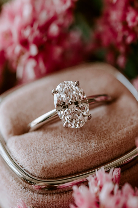 Oval Engagement Ring Obsession: Why This Cut Is Everyone's Main Character Right Now 8 Close-up of an oval diamond solitaire engagement ring on a blush velvet ring box with soft pink flowers in the background