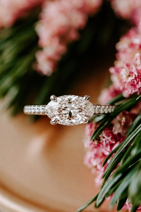 Oval Engagement Ring Obsession: Why This Cut Is Everyone's Main Character Right Now 10 Oval diamond solitaire engagement ring with pavé band nestled among soft pink flowers in warm natural light for a romantic look