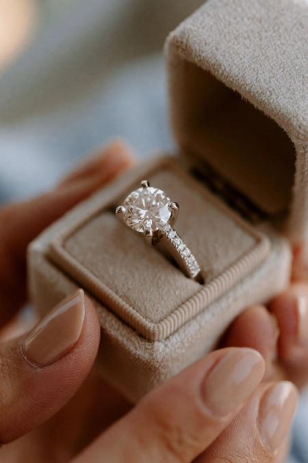 Proposal Ideas: 40 Ways to Pop the Question She'll Never Stop Talking About 12 Close-up of a round diamond engagement ring in an open velvet ring box held in hands with a soft neutral manicure