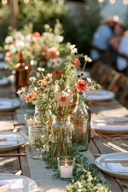 Rustic backyard rehearsal dinner centerpiece with wildflowers in glass jars and soft candlelight on wood tables