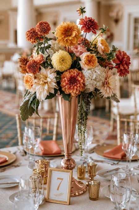 25 Fall Wedding Centerpieces With Main Character Energy (Cozy, Never Costume-y) 24 Tall copper vase centerpiece with caramel dahlias, roses, hydrangeas, and autumn blooms on an elegant wedding reception table