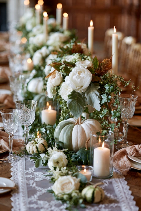 25 Fall Wedding Centerpieces With Main Character Energy (Cozy, Never Costume-y) 11 White pumpkin centerpiece with ivory roses, lush greenery, and pillar candles on an elegant fall wedding reception table