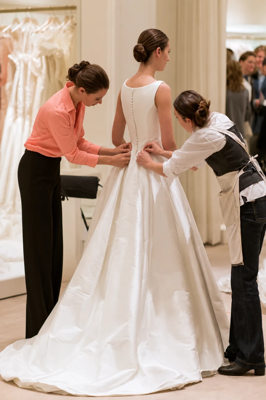 Bride in a white satin wedding gown at her fitting, with two seamstresses adjusting the waist and bodice in a bridal boutique